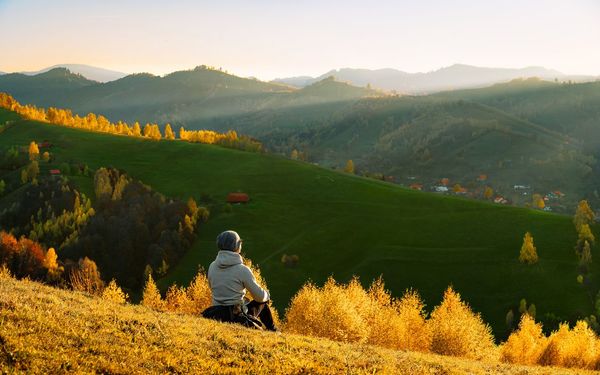 Rear view of man resting and looking at hills against mountains at sunset during autumn