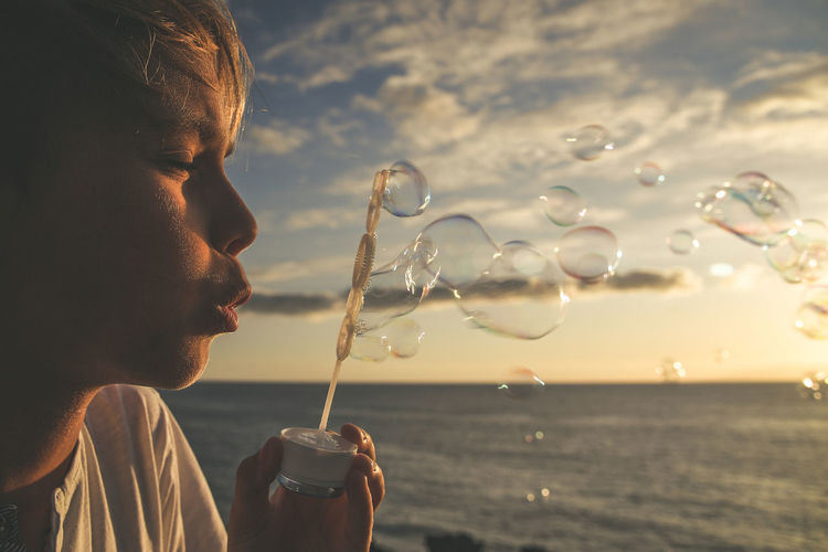 Close-up of cute boy blowing bubbles against sky during sunset