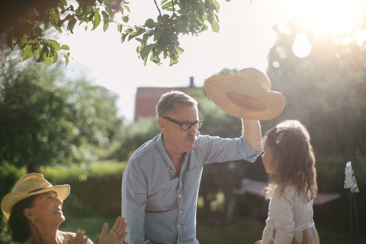 Woman clapping and looking at senior man holding straw hat over granddaughter in backyard during sunny day