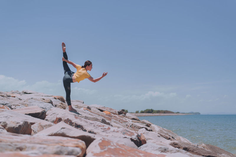 Woman exercising while standing on rock by sea against sky