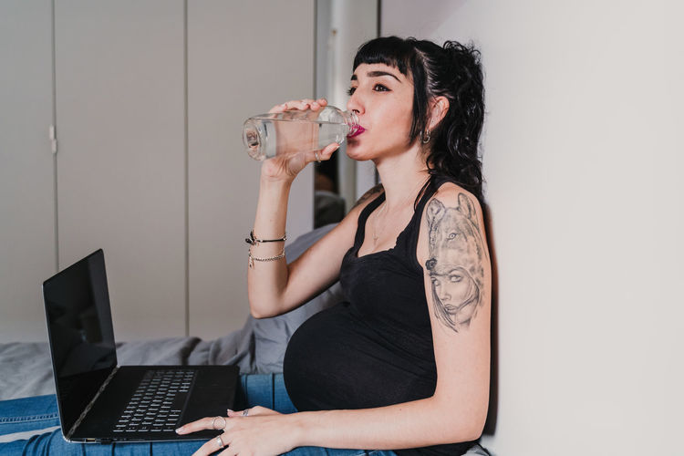 Pregnant young woman drinking water while sitting with laptop at home