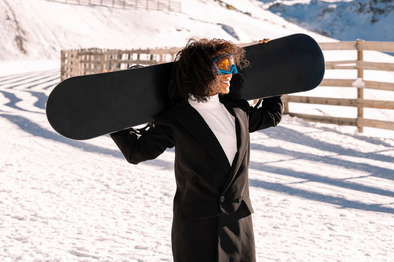 African american woman with goggles and a snowboard on a snowy mountain during winter