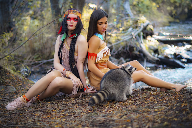 Young women in traditional clothing sitting by raccoon in forest