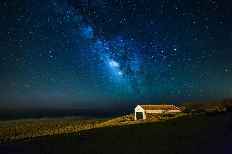 Building on sand by sea against milky way in sky at night