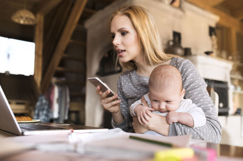 Mother with baby at home using laptop and cell phone