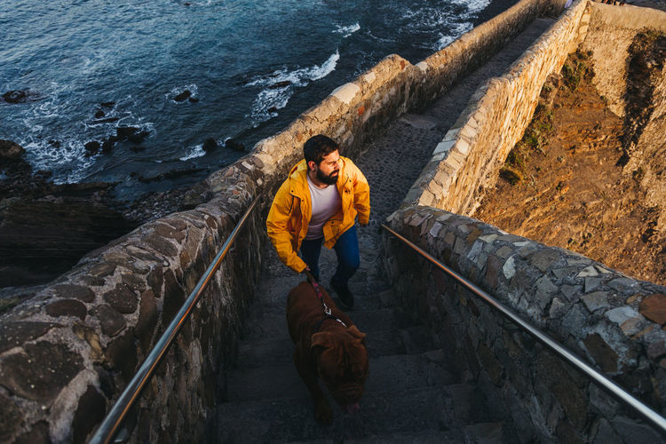 High angle of male in bright yellow jacket with big brown dog walking up stone stairs and looking away with interest against troubled bay water washing rocky coast in spain during sunrise