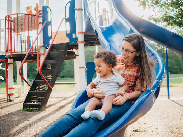 Smiling mother with cute kid sitting on slide at park