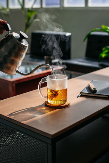 Crop anonymous person pouring hot tea from teapot into glass cup placed on wooden counter in room with bright sunlight person