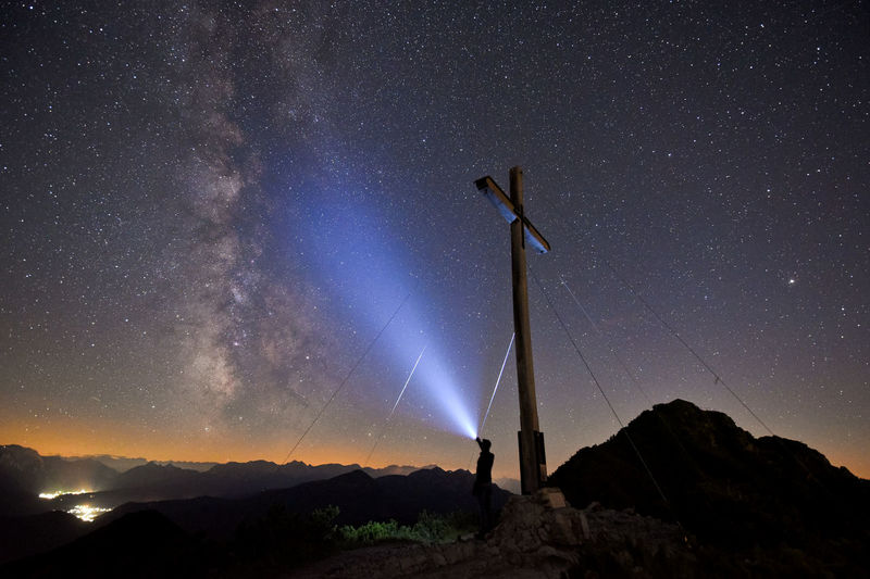 Man pointing flashlight towards star field while standing by cross on mountain peak at night