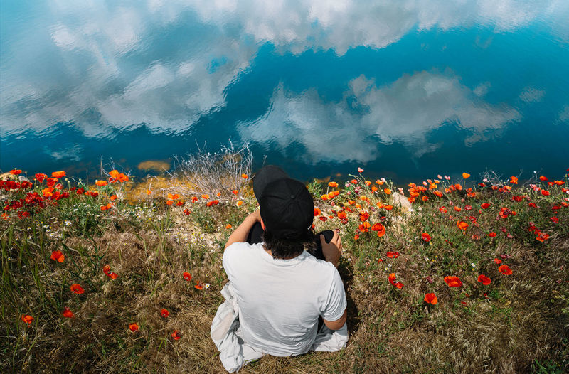 Man sitting in poppy field by the lake with clouds reflection