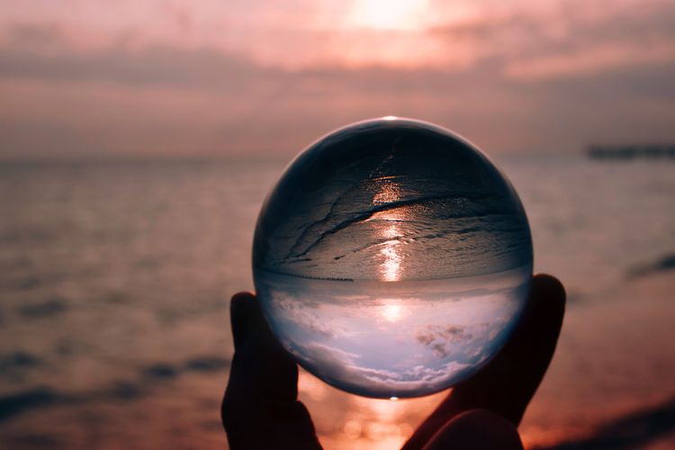 Close-up of hand holding crystal ball against sea during sunset