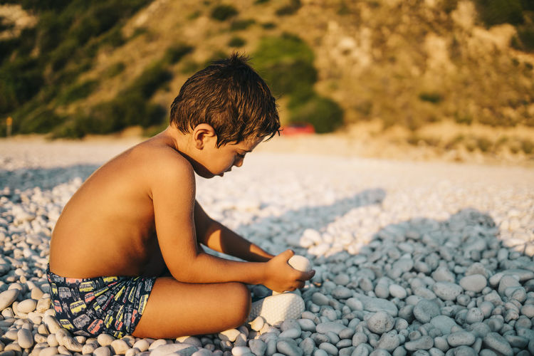 Side view of shirtless boy sitting on pebble