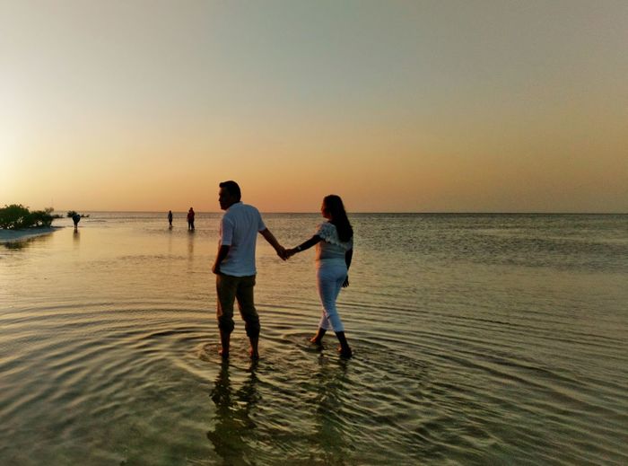 Rear view of couple holding hands while walking at beach during sunset