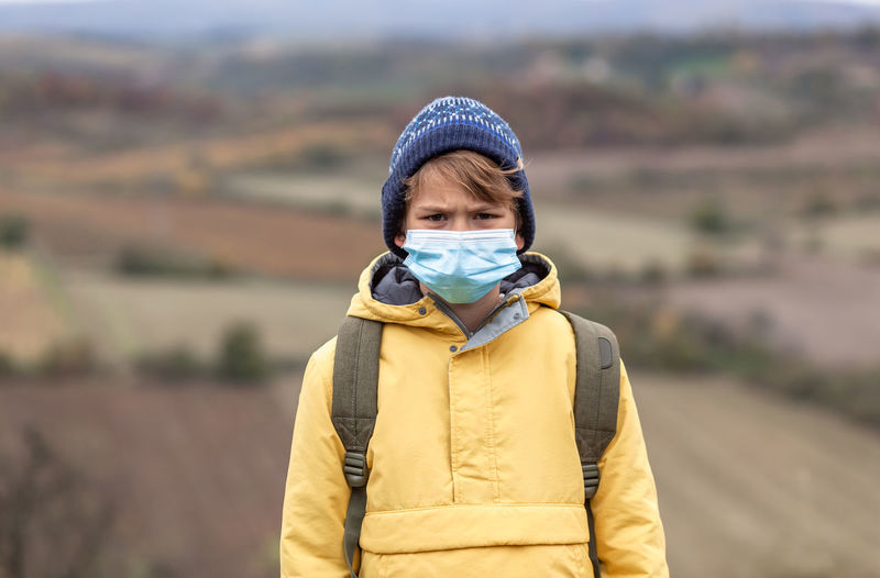 Portrait of a displeased little boy with face mask during pandemic in nature looking at camera