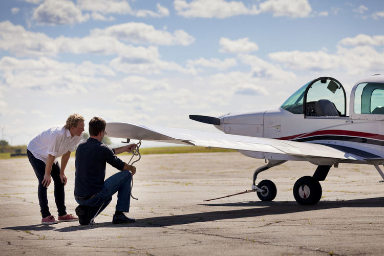 Mechanics repairing aircraft wing at airfield against cloudy sky