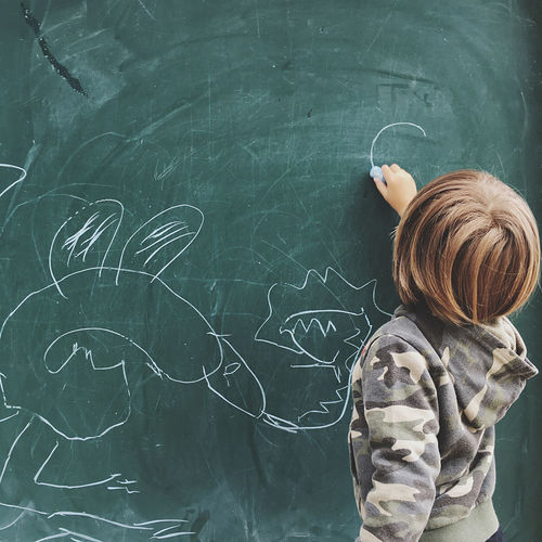 Rear view of boy drawing on blackboard at classroom
