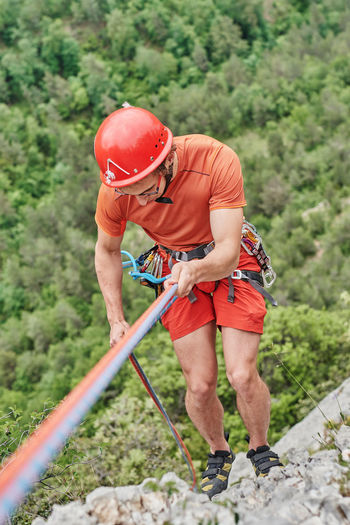 From above of brave male alpinist in protective equipment climbing steep cliff on background of forest