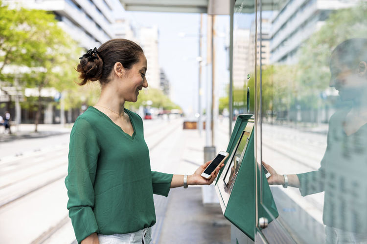 Happy woman using smart phone and ticket machine at tram stop