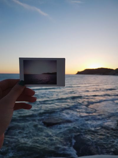 Cropped hand of woman holding instant print transfer against sea and sky during sunset