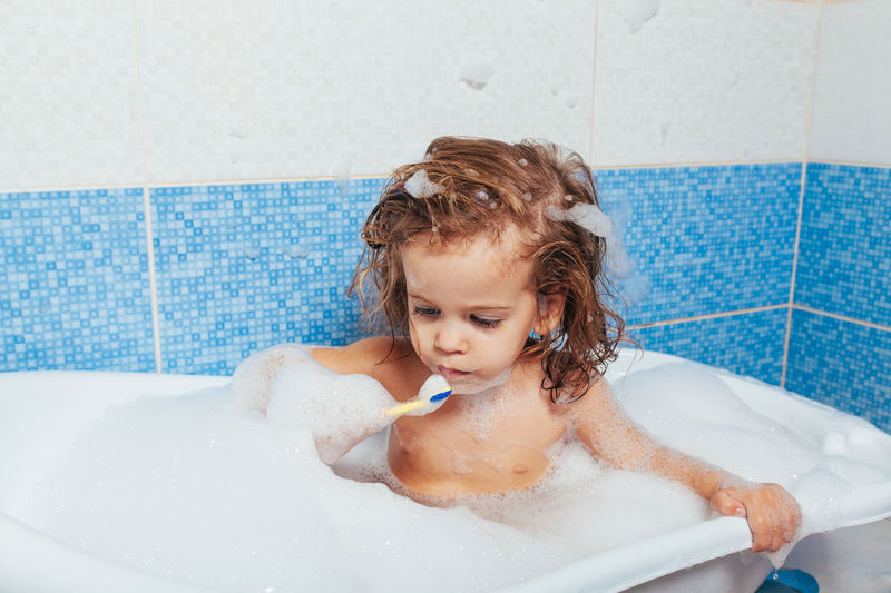 Topless girl brushing teeth while sitting in bathtub at home