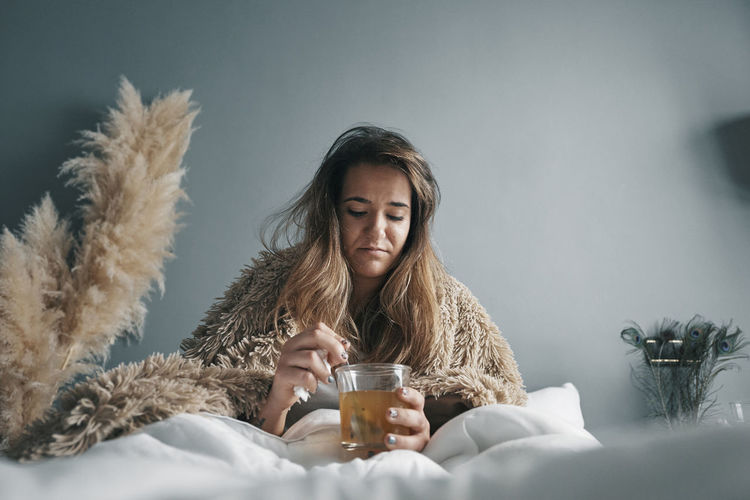 Young woman with herbal tea sitting on bed against wall