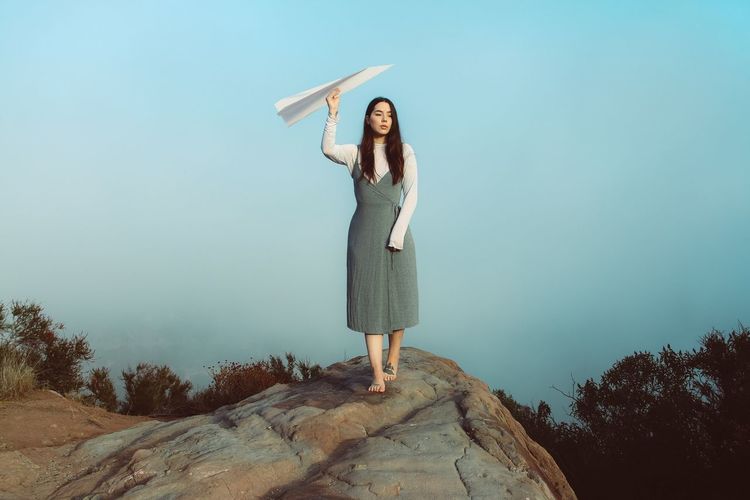 Woman holding paper airplane while walking on rock against sky during sunset