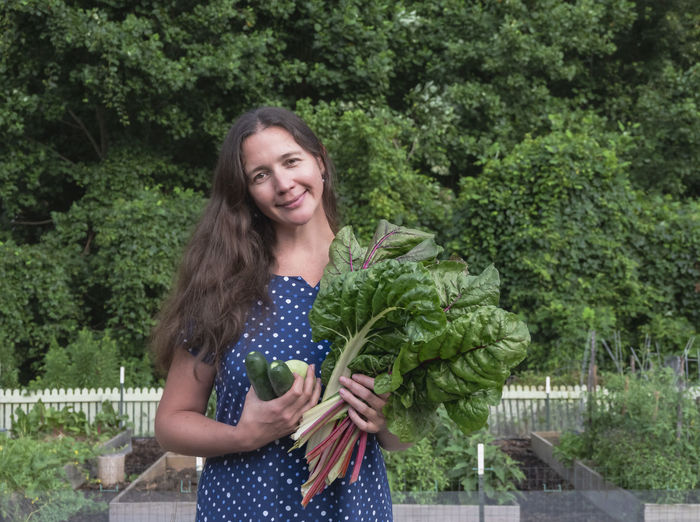 Portrait of smiling woman holding vegetable standing against plants