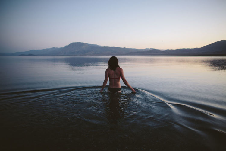 Rear view of woman wearing bikini while standing in lake against clear sky during dusk