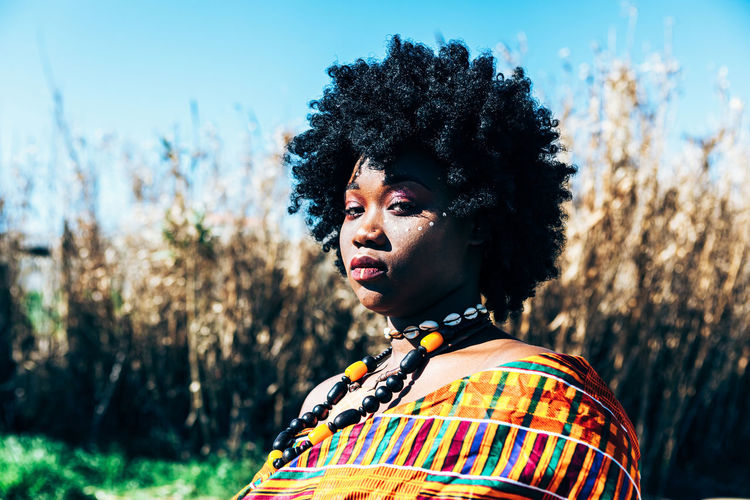 Serious african model with afro hairstyle wearing colorful handwoven kente and looking at camera while standing on field on bright summer day