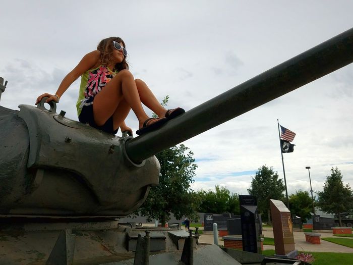 Full length of girl sitting on armored tank at war memorial against sky