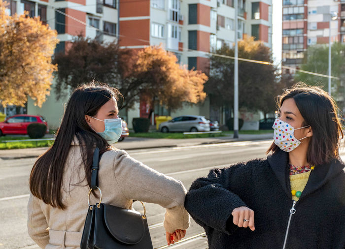 Two women walking in city, wearing masks during corona virus epidemic, doing elbow bump handshake