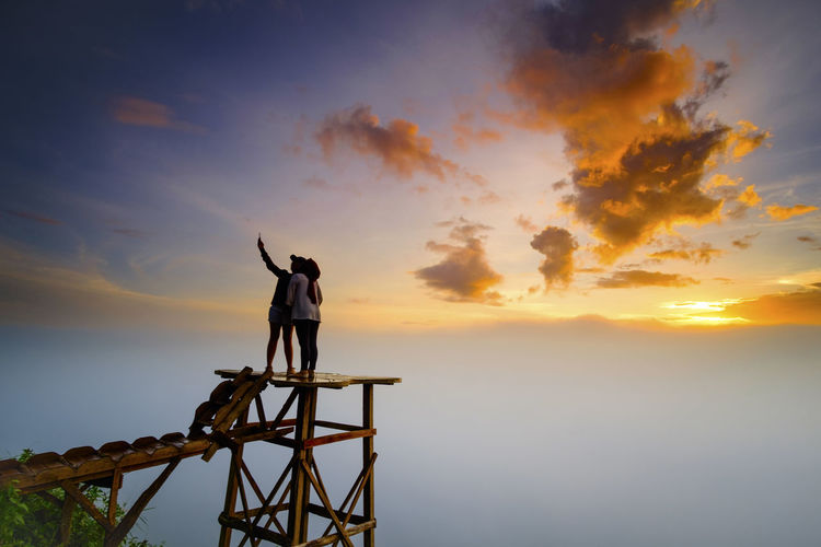 Silhouette couple taking selfie while standing on observation point against cloudy sky during sunset