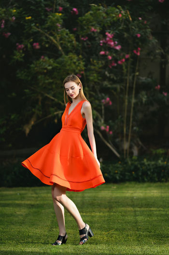Full length of young woman in orange dress standing on grassy field at park