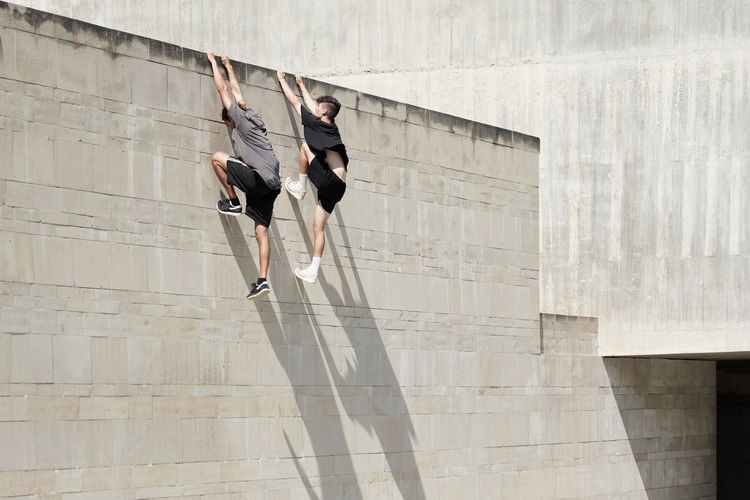 Side view of brave male friends hanging on wall of concrete building while performing risky trick and doing parkour in city