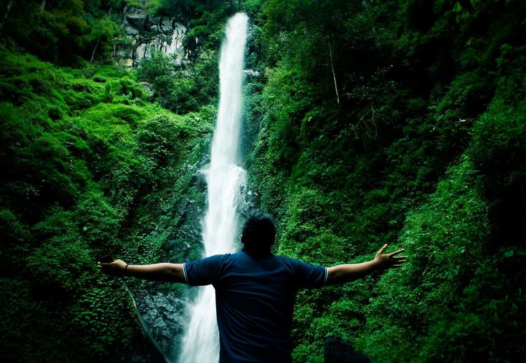 Rear view of man with arms outstretched against waterfall in forest