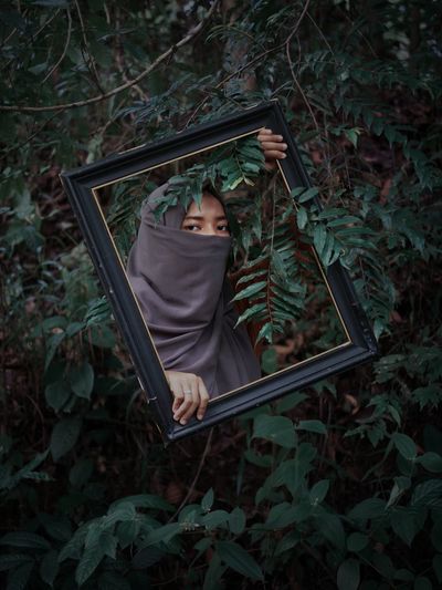 Portrait of woman wearing hijab holding picture frame against plants