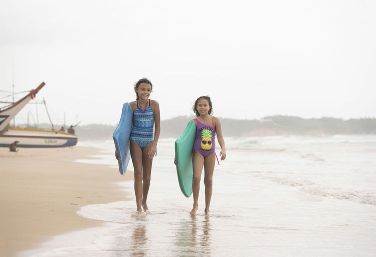 Full length of sisters with surfboards walking on shore at beach against sky