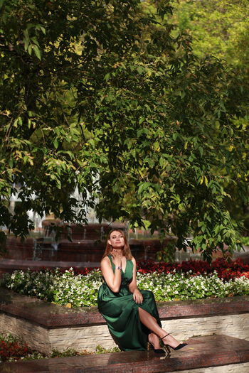 Portrait of woman sitting by tree against autumn park background, wearing green silk dress 