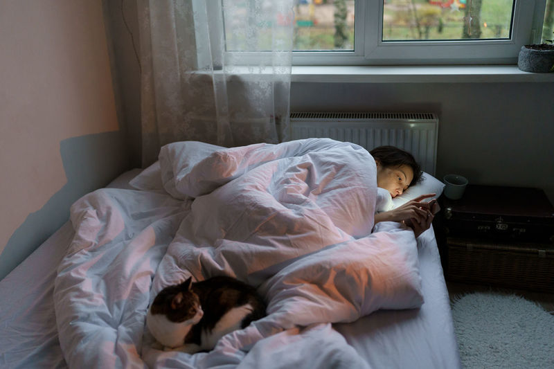 Young woman addicted to social media and internet using mobile, lonely female lying in bed with cat