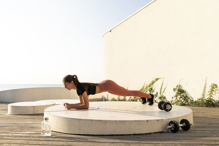 Sportswoman doing plank position exercise on pedestal by sea during sunrise