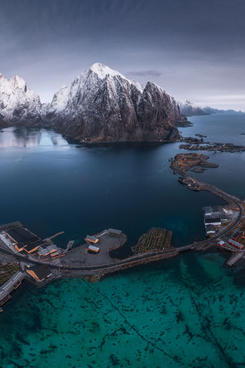 From above aerial view of calm water of sea washing island with a village and rocky formations covered in snow under cloudy sky in uttakleiv beach in lofoten, norway