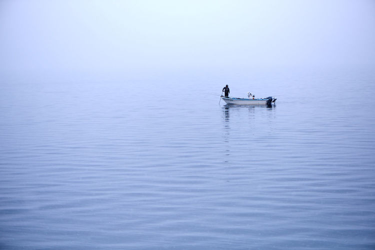 Man standing on moored fishing boat on calm lake during foggy weather