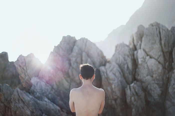 Rear view of shirtless man standing by rocks against sky