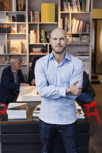 Portrait of confident businessman standing arms crossed while colleagues working in background at office