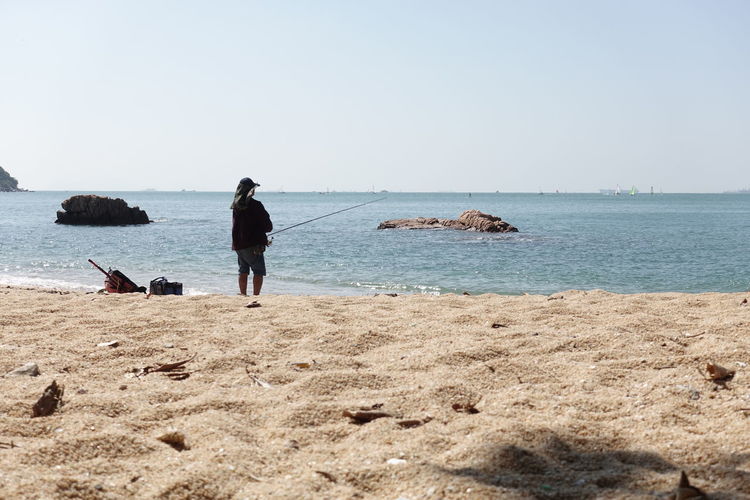 Rear view of man fishing on beach against clear sky