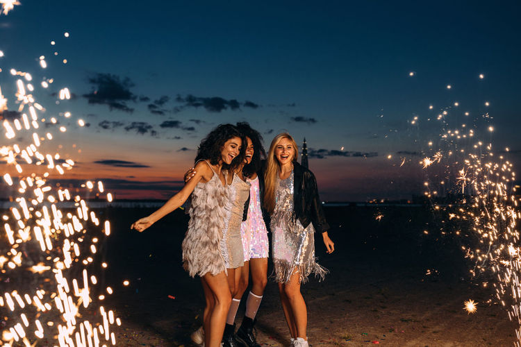 Cheerful young female friends with firework at beach against sky during sunset