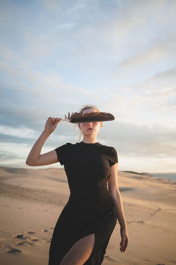 Serious female covering her eyes with black feather wearing dress standing on sandy dune washed by sea at sundown