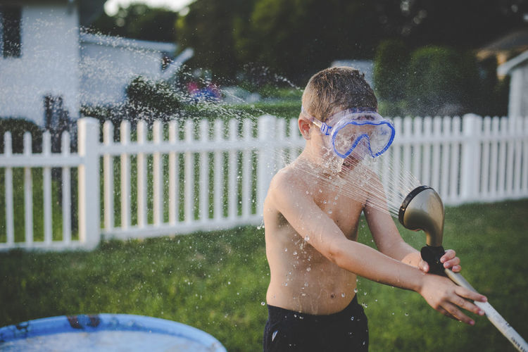 Playful shirtless boy wearing swimming goggles while spraying hose on him at yard