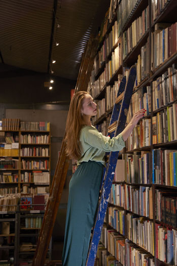 Woman with long hair standing on ladder while searching book in library
