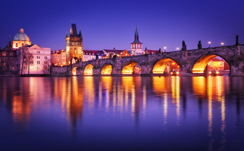 Charles bridge over river by buildings in illuminated city against clear sky